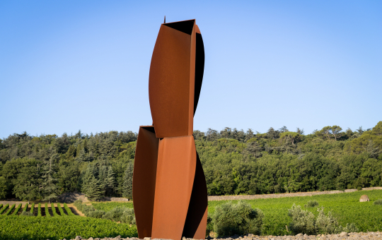 Sculpture en métal rouillé dans un paysage verdoyant sous un ciel bleu.