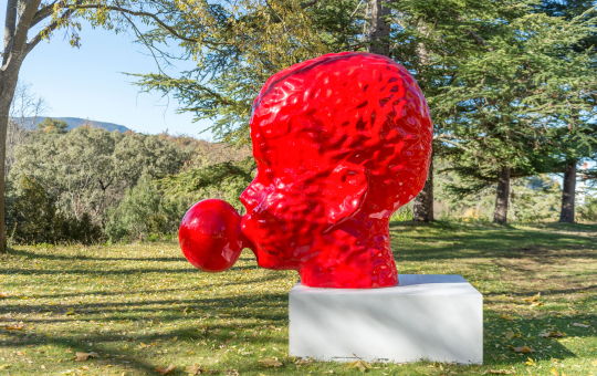 Sculpture rouge sur socle blanc dans un parc ensoleillé, œuvre d'art moderne.