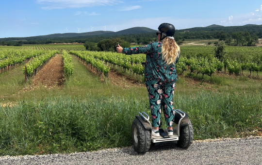 Une femme sur gyropode explore un vignoble ensoleillé avec un geste de la main.