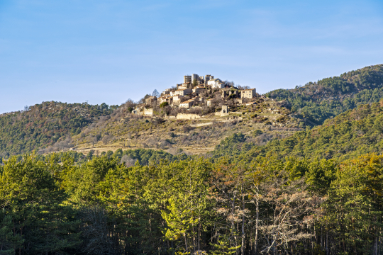 Un village médiéval perché sur une colline, entouré de forêts verdoyantes sous un ciel bleu.