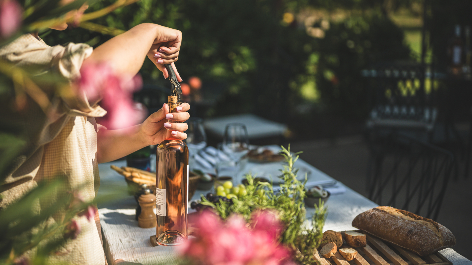 Une femme ouvre une bouteille de rosé sur une table en plein air, avec des fleurs et du pain.