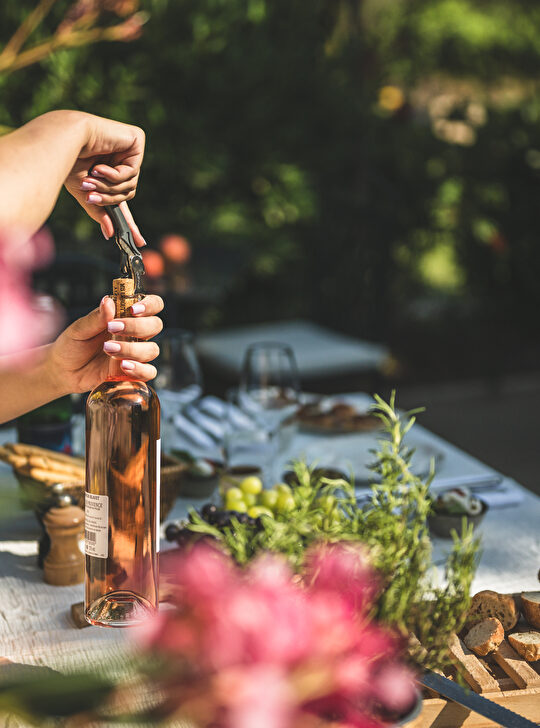 Une femme ouvre une bouteille de rosé sur une table en plein air, avec des fleurs et du pain.