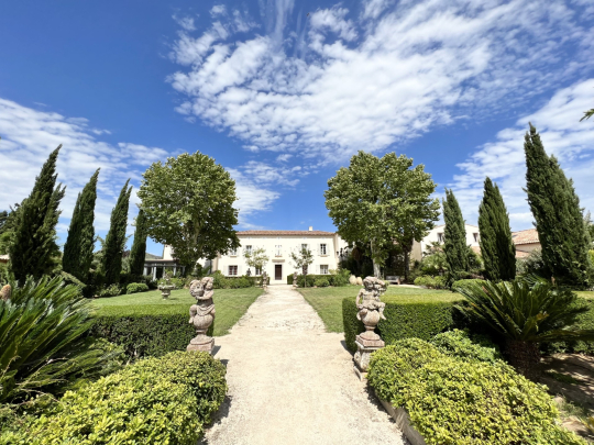 Un beau manoir entouré d'un jardin luxuriant et d'allées élégantes sous un ciel bleu.