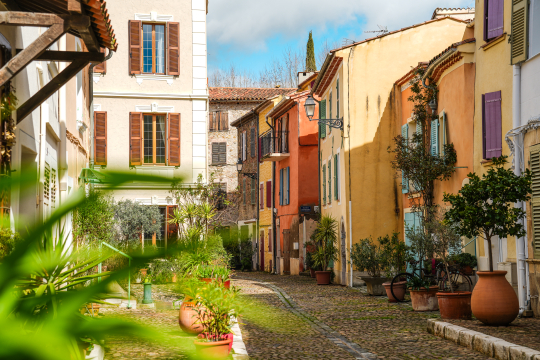 Une rue pittoresque colorée dans une ville méditerranéenne, avec des bâtiments anciens et des pots de fleurs.