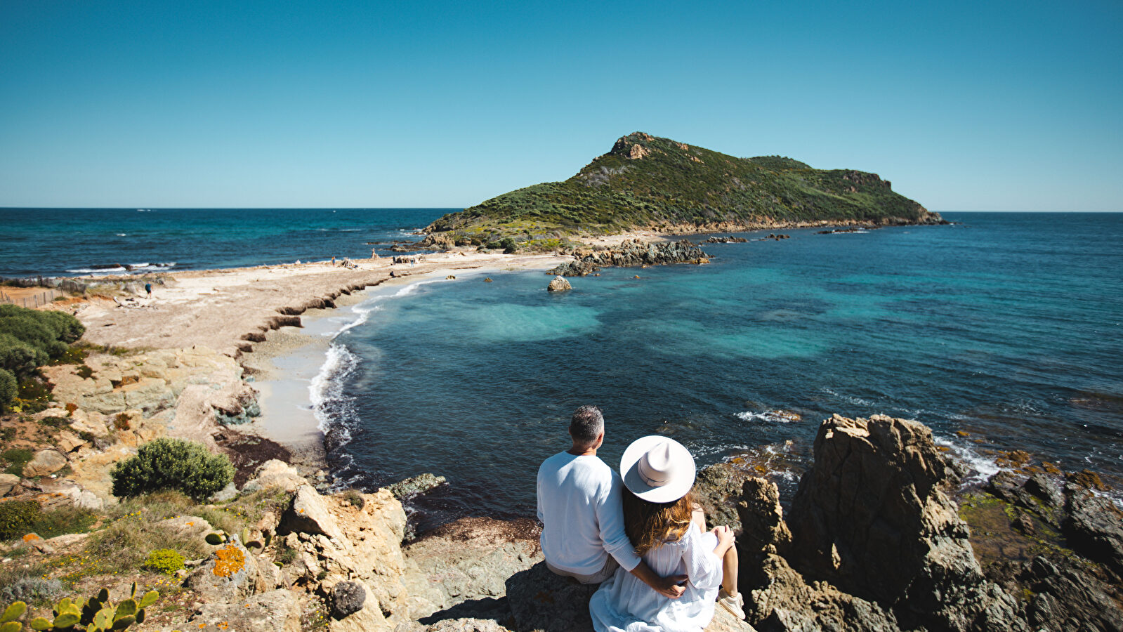 Un couple admire une côte méditerranéenne pittoresque avec une île au loin.