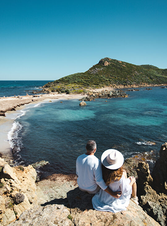 Un couple admire une côte méditerranéenne pittoresque avec une île au loin.