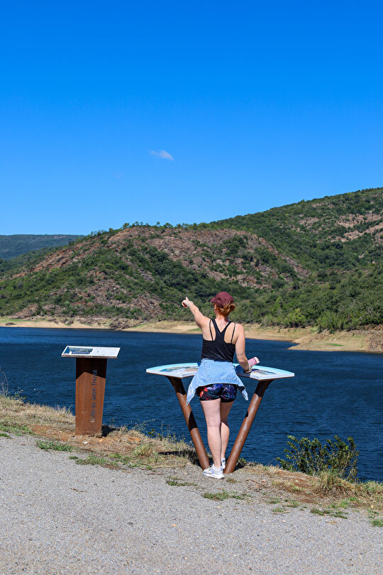 Une femme admire le paysage lacustre et les collines verdoyantes sous un ciel bleu éclatant.